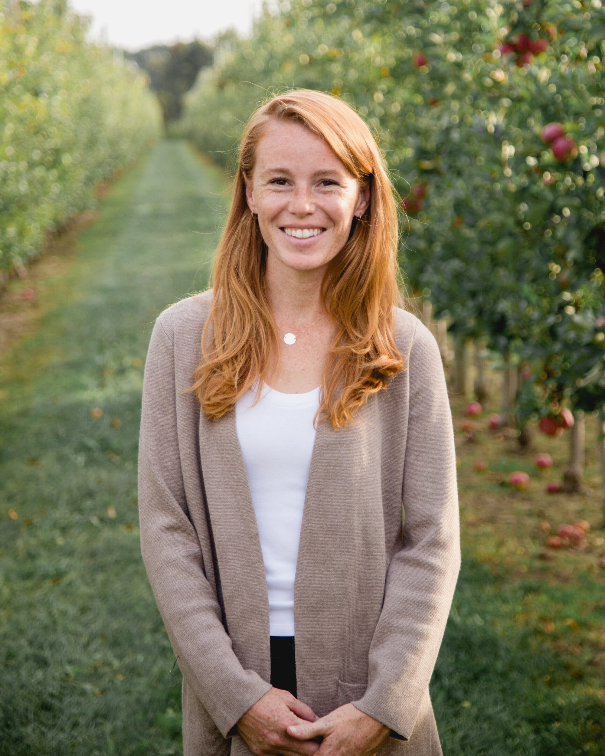 A picture of Democratic nominee for Wisconsin's Third District Rebecca Cooke. She is a woman in her 30s with red hair, standing in the middle of an apple orchard, smiling with her hands clasped together.